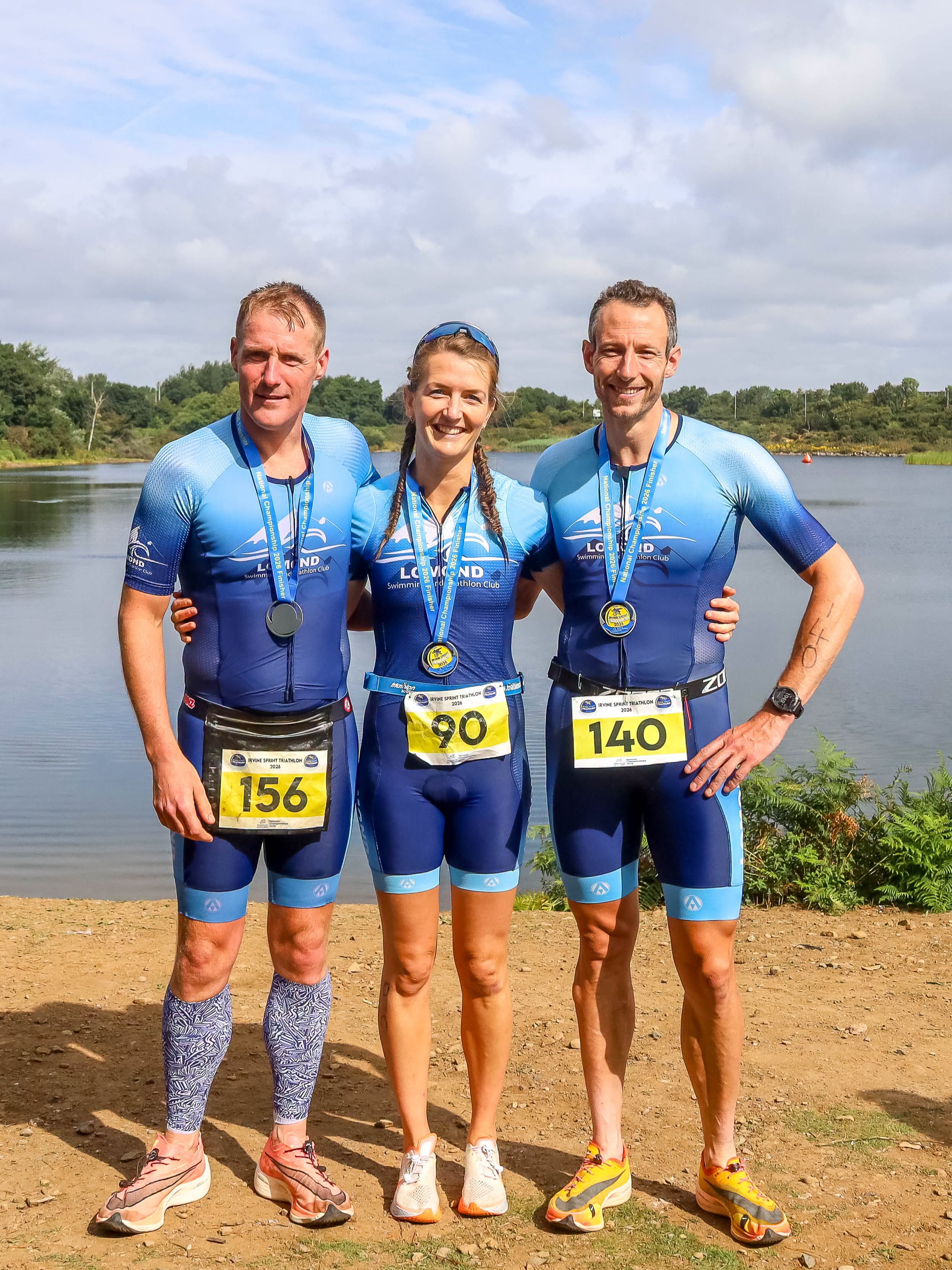 Three Lomond members in club kit with their finishers' medals beside the water